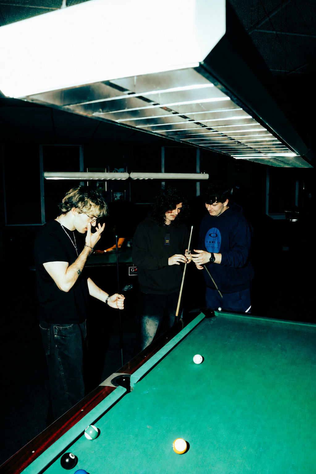 Three men standing around a green pool table in a dimly lit room, with one holding a pool cue and two others observing. Overhead fluorescent lighting creates a bright glare near the top of the frame.